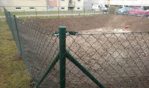 Maschendrahtzaun mit grünem Metallrahmen und einem stabilen Pfostensystem, das an einem Erdgrundstück montiert ist. Der Zaun grenzt an eine leere Fläche, und im Hintergrund sind Wohngebäude sichtbar.