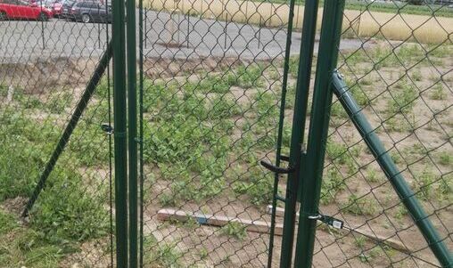 Doppeltes Metalltor mit grünem Maschendraht, ausgestattet mit einer mittigen Verschlussvorrichtung. Die Pfosten sind mit diagonalen Verstrebungen stabilisiert. Im Hintergrund sind ein unebenes Grundstück und ein geparktes Fahrzeug erkennbar.