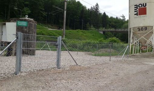 Verzinktes Metalltor mit senkrechten Gitterstäben, das Teil eines langen Maschendrahtzauns ist. Der Zaun umschließt ein industrielles Gelände mit einem Kiesweg und einem kleinen Gebäude im Hintergrund.