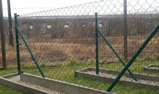 Maschendrahtzaun mit grün beschichteten Metallpfosten, die durch diagonal verlaufende Verstrebungen stabilisiert sind. Der Zaun ist auf einem Betonsockel montiert und befindet sich auf einem unbebauten Grundstück mit trockenem Gras und industriellem Hintergrund.