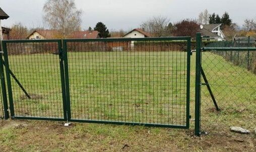 Grünes Metalltor mit integriertem Maschendrahtzaun, montiert auf einem ebenen Rasenstück. Das Tor ist an einem stabilen Metallrahmen befestigt, und im Hintergrund sind Wohnhäuser und Bäume sichtbar.