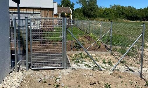 Kombination aus einem stabilen Maschendrahtzaun und einem verzinkten Metalltor mit vertikalen Gitterstäben. Der Zaun grenzt an ein bepflanztes Grundstück, während das Tor einen Zugang zu einem gepflasterten Bereich bietet.