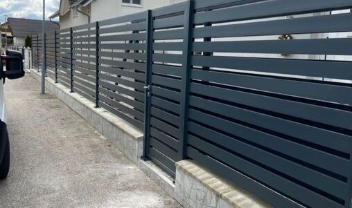 Langer Aluminiumlattenzaun in elegantem Design, montiert auf einer niedrigen Betonsockelwand, entlang einer Straße in einem Wohngebiet.