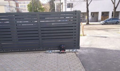 Dunkles, modernes Schiebetor aus horizontal angeordneten Aluminiumlatten, montiert auf einer asphaltierten Einfahrt, mit Blick auf ein Wohnviertel im Hintergrund.