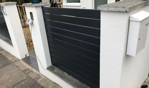 Schlichtes Gartentor in dunklem Aluminiumdesign, integriert in eine weiße Betonsockelwand, mit einem Briefkasten und einer Klingelanlage daneben.