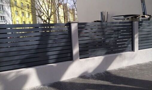 Eleganter Zaun aus dunkelgrauen Aluminiumlatten, montiert auf einer niedrigen weißen Betonsockelwand. Der Zaun umschließt einen gepflasterten Hof mit Schattenwurf.