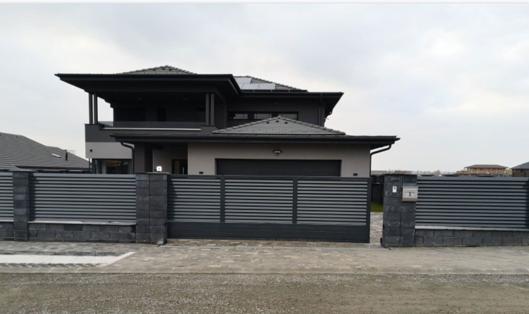 Moderner Zaun mit waagerechten grauen Lamellen, der ein Wohnhaus umrahmt, ergänzt durch einen Steinunterbau.
