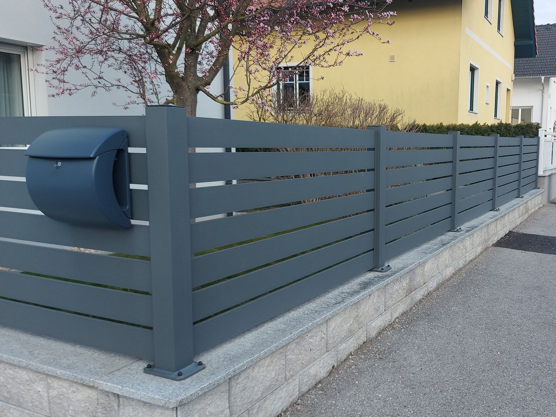 Grauer Metallzaun mit horizontalen Lamellen vor einem Wohnbereich, ergänzt durch ein bläulich-graues Briefkastenmodul.