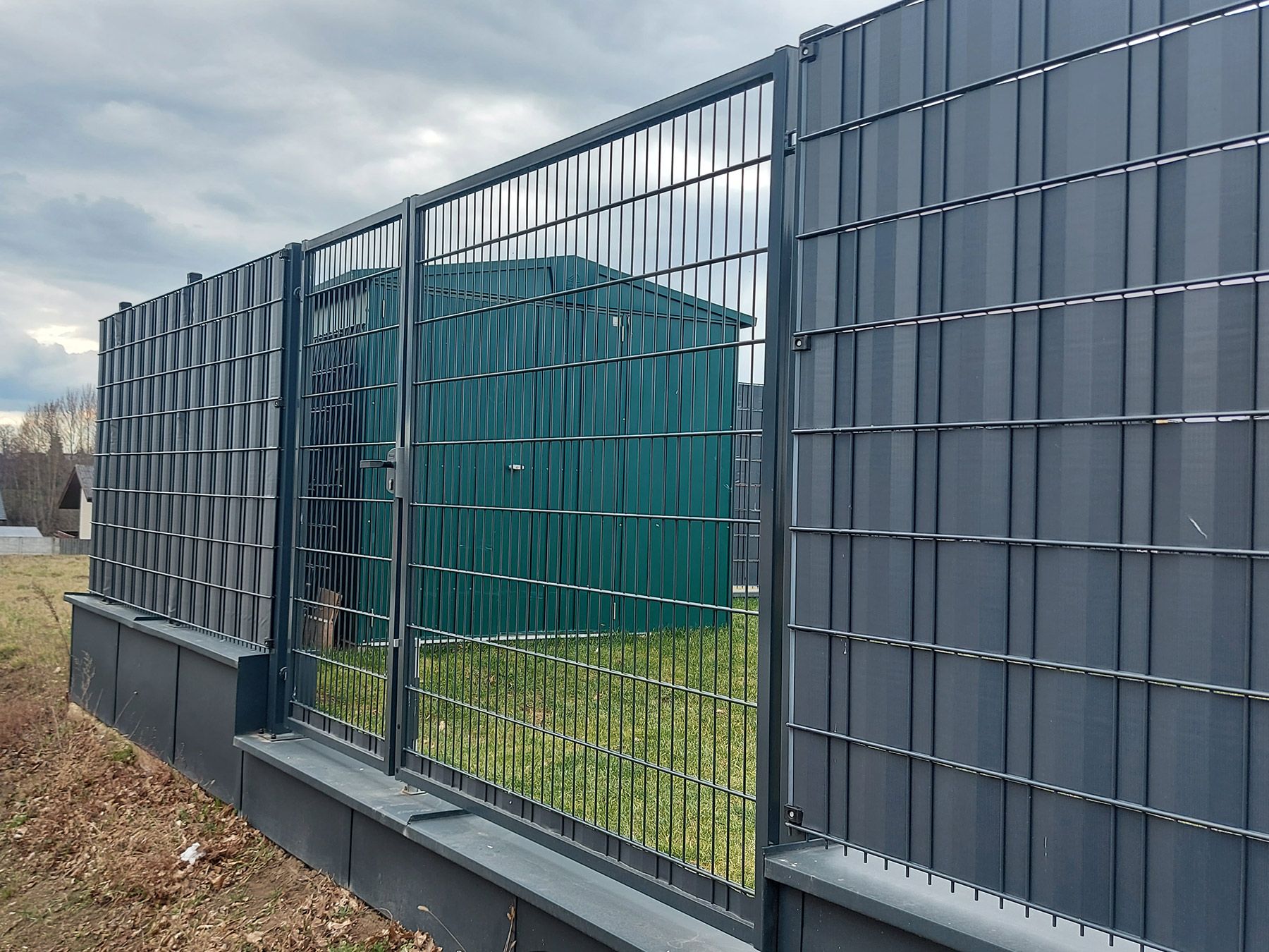 Metallzaun mit Gitterelementen, hinter dem ein grünes Gartenhaus zu sehen ist, mit einer verschlossenen Tür.