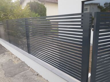 Zaun mit schwarzen Lamellen und Schiebetür, der einen Wohnbereich abschließt, umgeben von Gartenflächen.